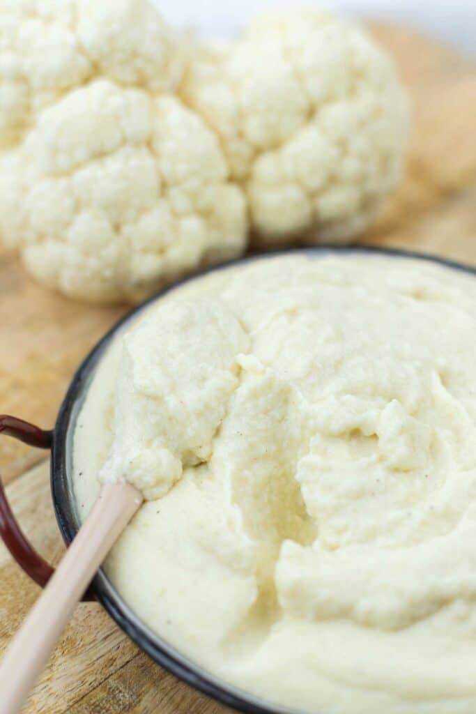 A bowl of creamy mashed cauliflower with a smooth texture, served alongside fresh cauliflower heads on a wooden surface.