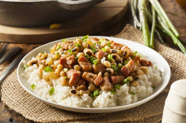 A plate of Southern Hoppin' John, featuring fluffy white rice topped with black-eyed peas, diced ham, and chopped green onions, served on a rustic burlap mat with fresh green onions in the background.