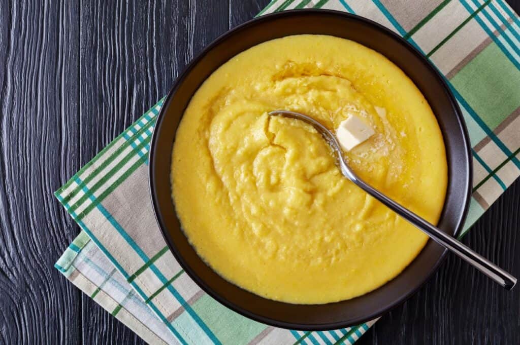 Hot cornmeal polenta with melting butter in a bowl with napkin and silver spoon on a black wooden table, horizontal view from above.