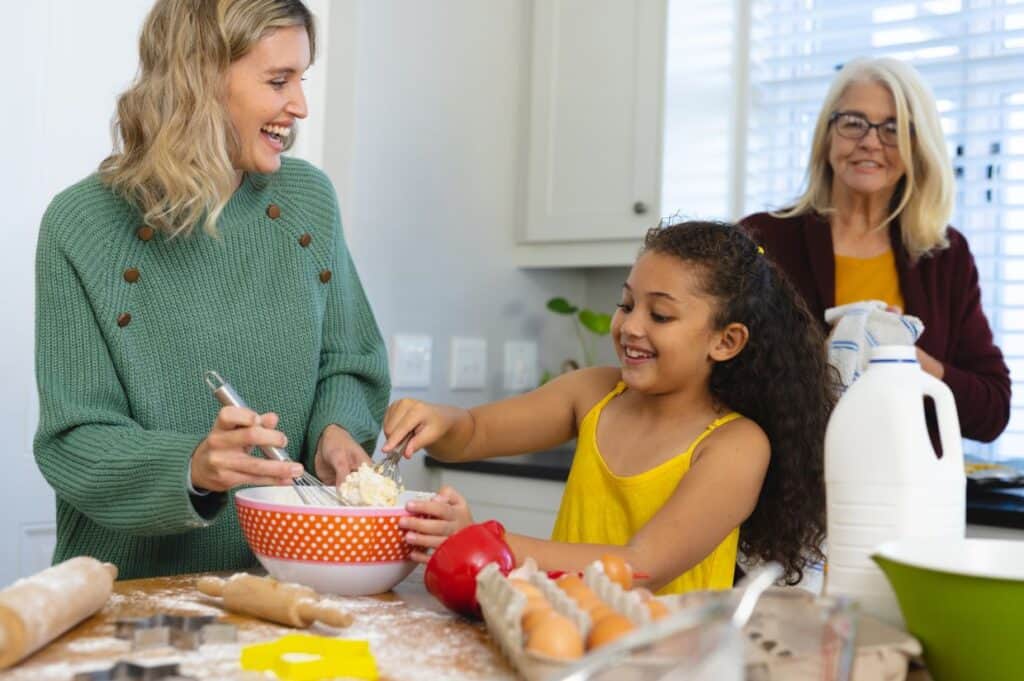 Multiracial senior woman looking at happy daughter and granddaughter mixing food in bowl in kitchen.