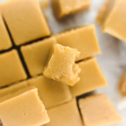 A stack of creamy old-fashioned peanut butter fudge cut into small squares, placed on parchment paper, with one piece partially bitten and an unwrapped piece in the foreground.