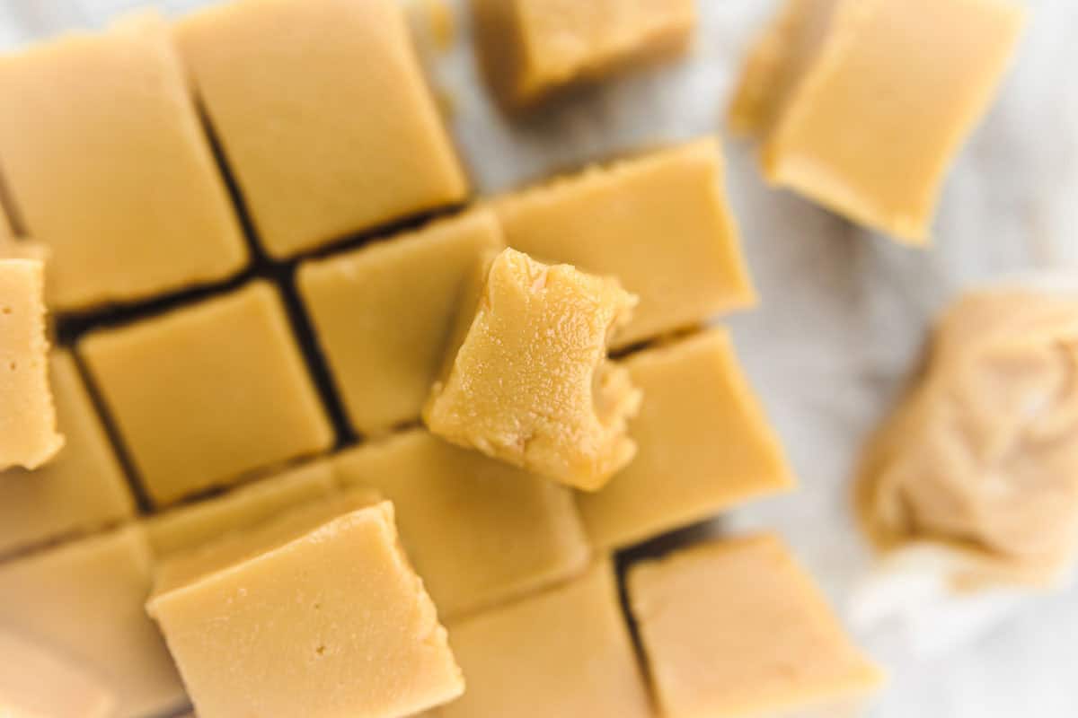 A stack of creamy old-fashioned peanut butter fudge cut into small squares, placed on parchment paper, with one piece partially bitten and an unwrapped piece in the foreground.