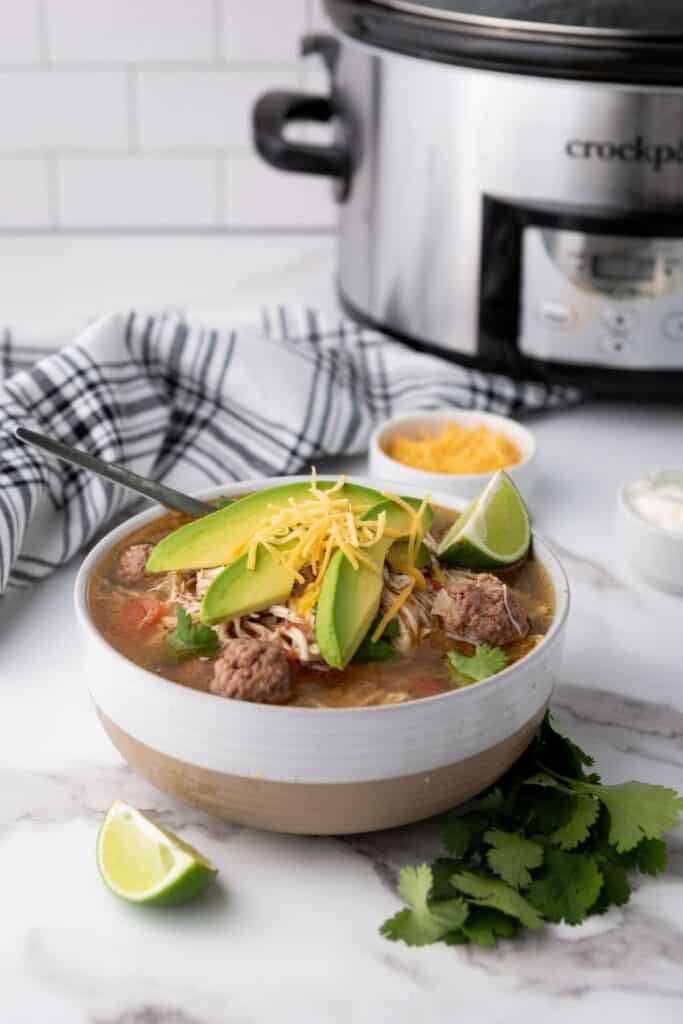 A bowl of taco soup topped with sliced avocado, shredded cheese, and a lime wedge.