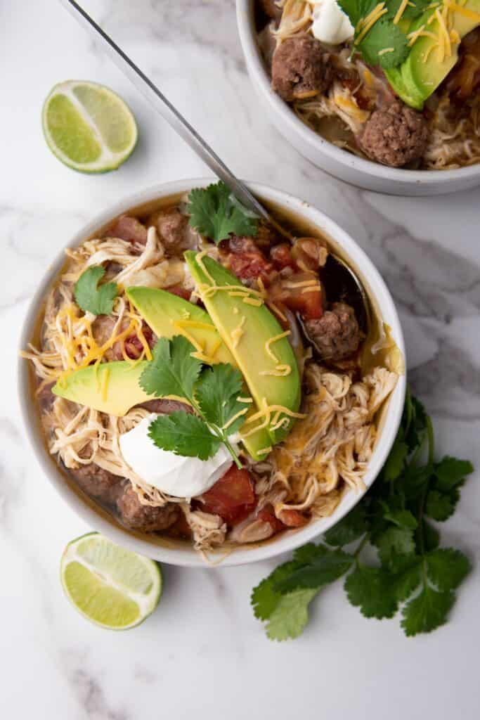 A bowl of taco soup with shredded cheese, avocado, and a spoon resting inside.