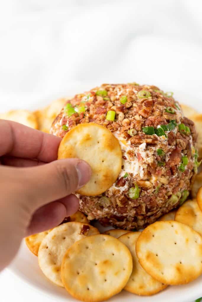 Close-up of a savory cheeseball coated with bacon, pecans, and green onions, paired with crackers.