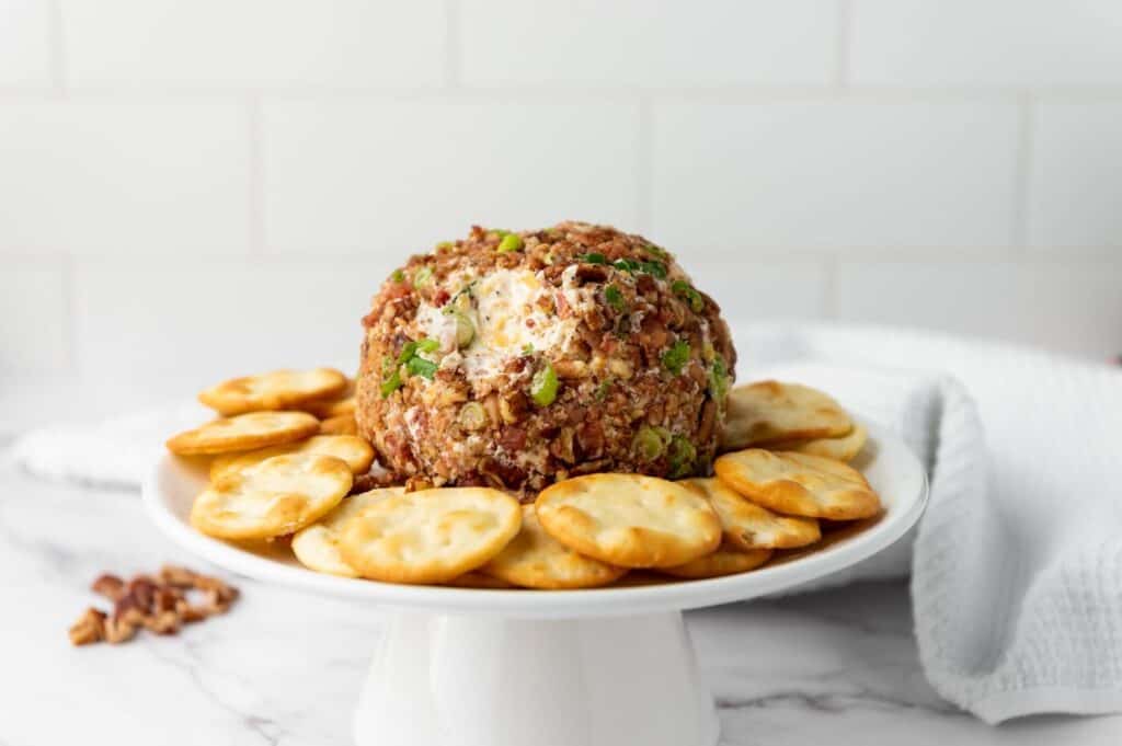 A pecan bacon cheeseball served on a white pedestal platter surrounded by crackers.