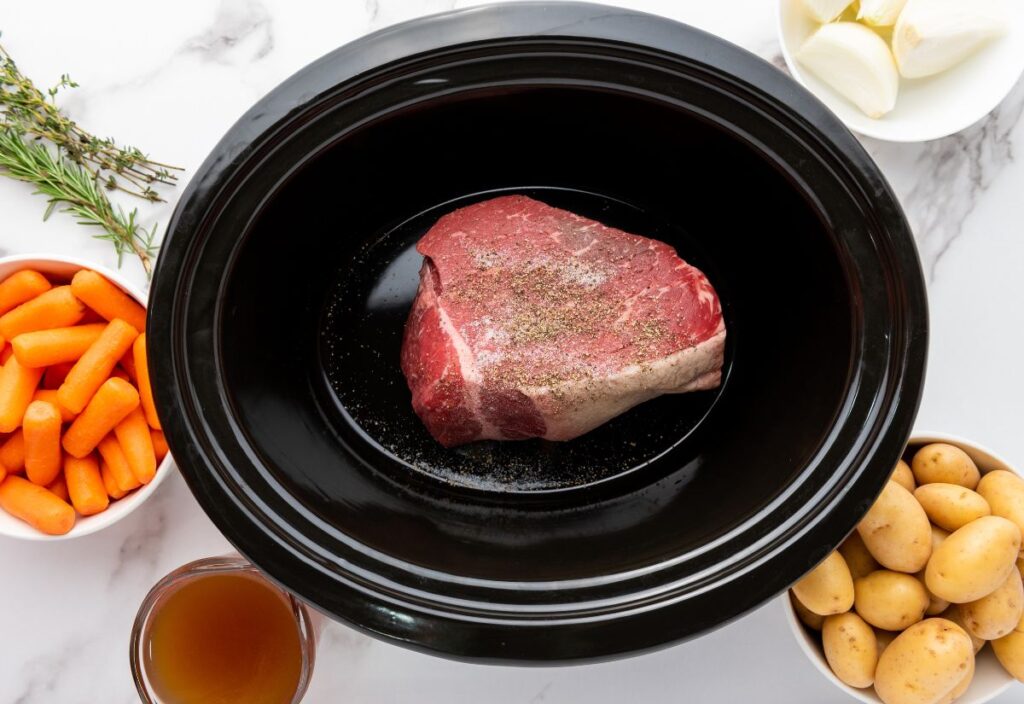 Raw beef roast in slow cooker with carrots, potatoes, onions, herbs, and broth.