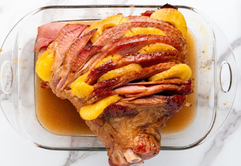 Baked ham with pineapple slices, glazed in a glass baking dish, sitting in juices.