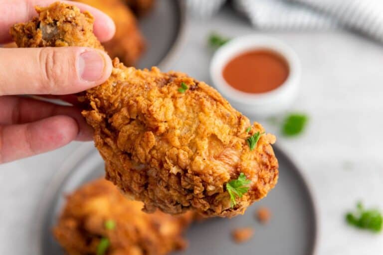 A close-up of crispy buttermilk fried chicken being held in hand, with a small bowl of dipping sauce and garnished parsley in the background.