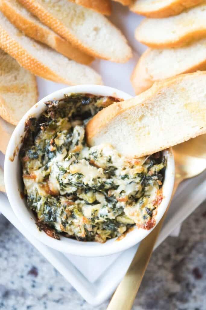 A cheesy, baked collard dip served in a white ramekin, with a crispy toasted bread slice partially dipped. 