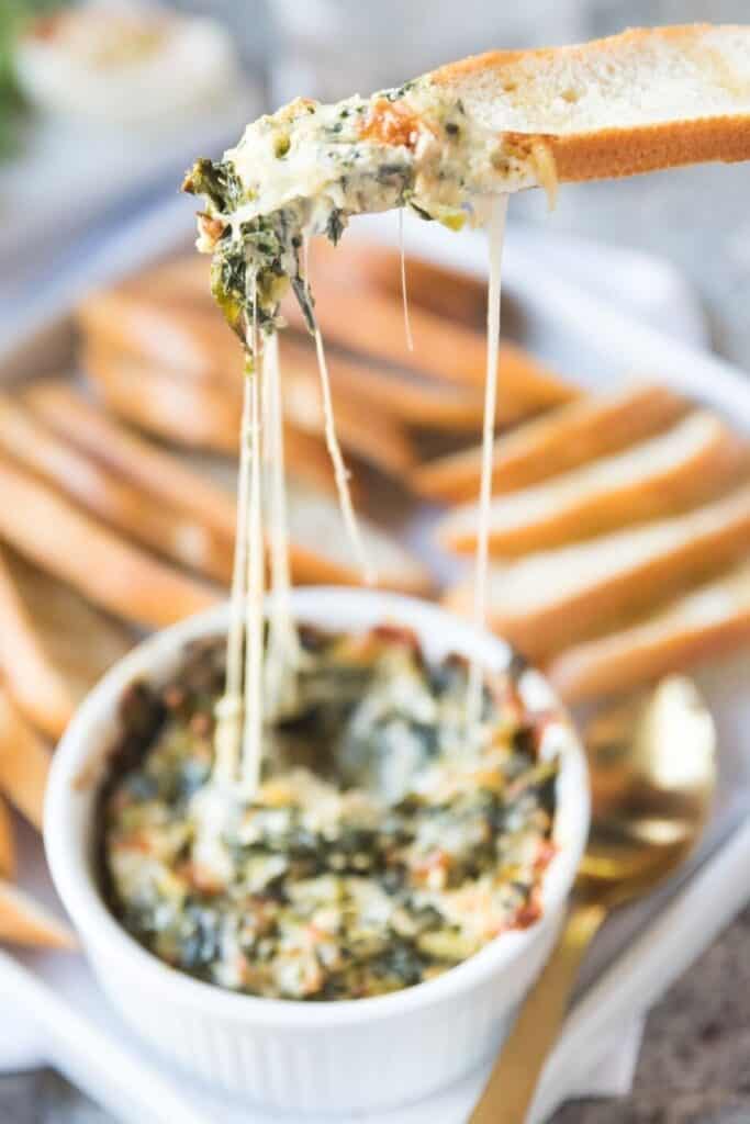 A gooey, cheesy collard dip served in a white ramekin, with melted cheese stretching as a piece of toasted bread is lifted.