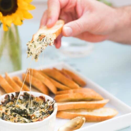 A creamy, cheesy collard dip served in a white ramekin, with melted cheese stretching as someone scoops the dip onto a crispy bread slice.