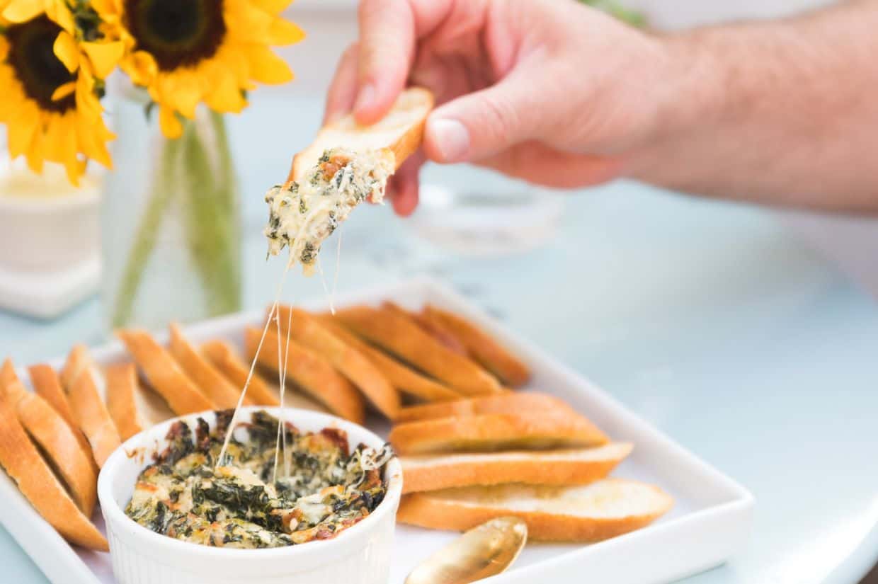 A creamy, cheesy collard dip served in a white ramekin, with melted cheese stretching as someone scoops the dip onto a crispy bread slice.