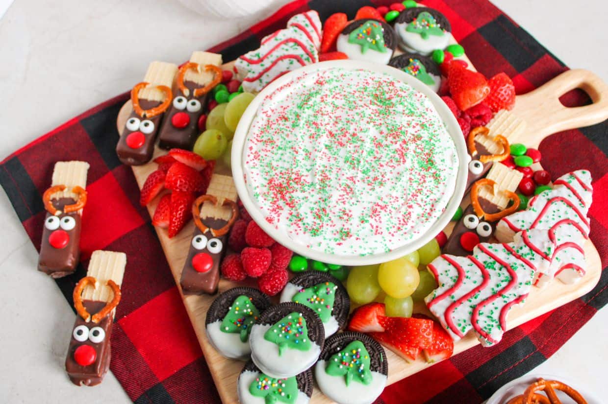 A festive Christmas charcuterie board with a bowl of sprinkled fruit dip surrounded by holiday-themed treats and fruits.