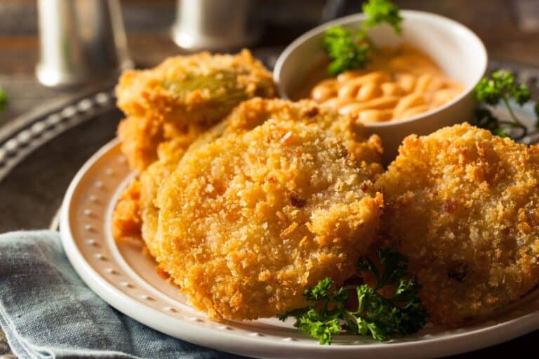 Plate of crispy fried green tomatoes coated in golden breadcrumbs, served with a side of dipping sauce and garnished with fresh parsley.