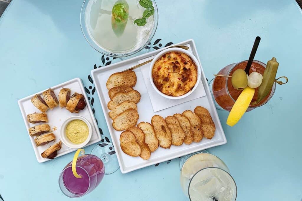 A table setting featuring a tray with hot crab and pimento cheese dip surrounded by toasted bread slices, a small plate of sausage rolls with mustard dip, and various colorful cocktails, including a Bloody Mary garnished with vegetables and a lemon wedge.