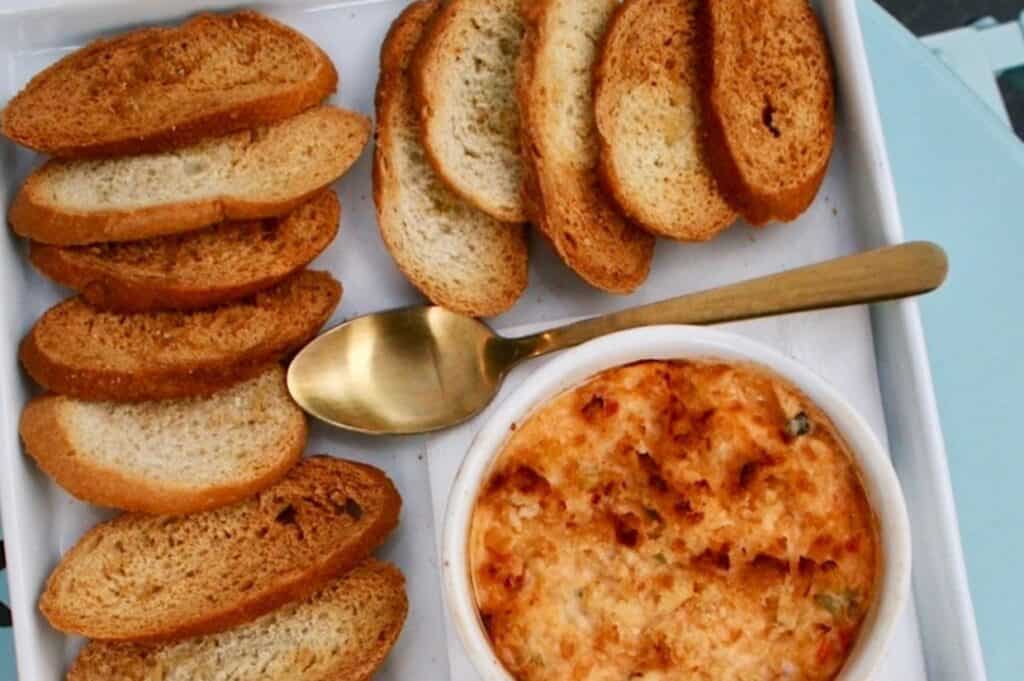 A bowl of hot crab and pimento cheese dip with a golden, bubbly surface is placed on a tray.