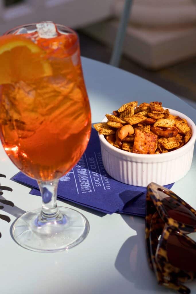 A close-up of a vibrant orange cocktail garnished with an orange slice, next to a white bowl filled with Chex Mix snacks on a blue napkin.