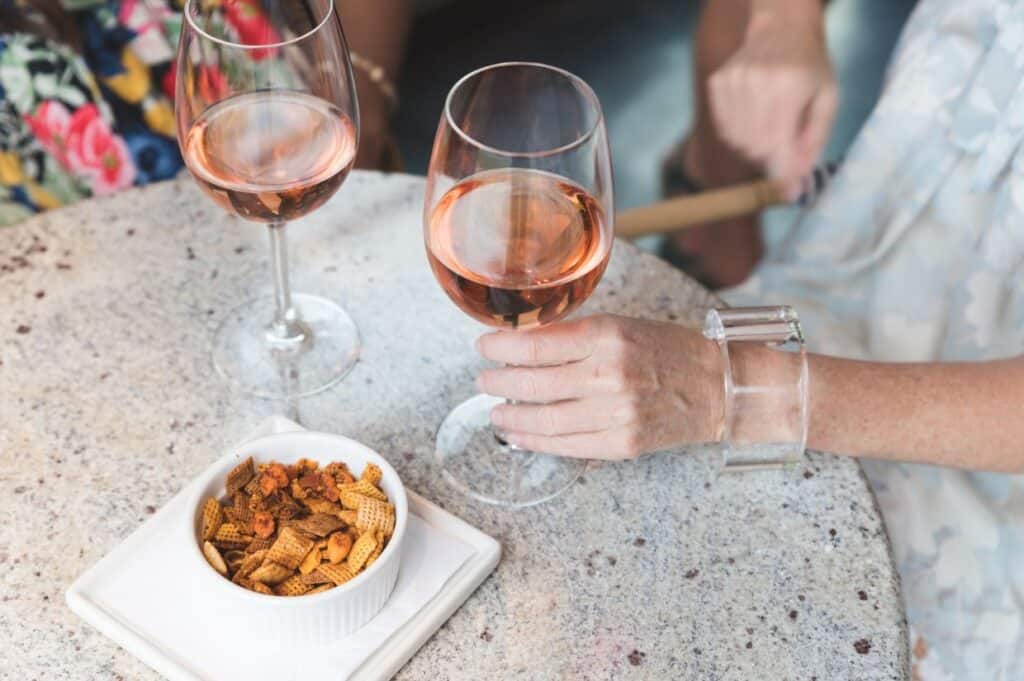 A close-up of two wine glasses filled with rosé on a speckled tabletop, accompanied by a white bowl of Chex Mix snacks. A hand with a clear bracelet is holding one wine glass.