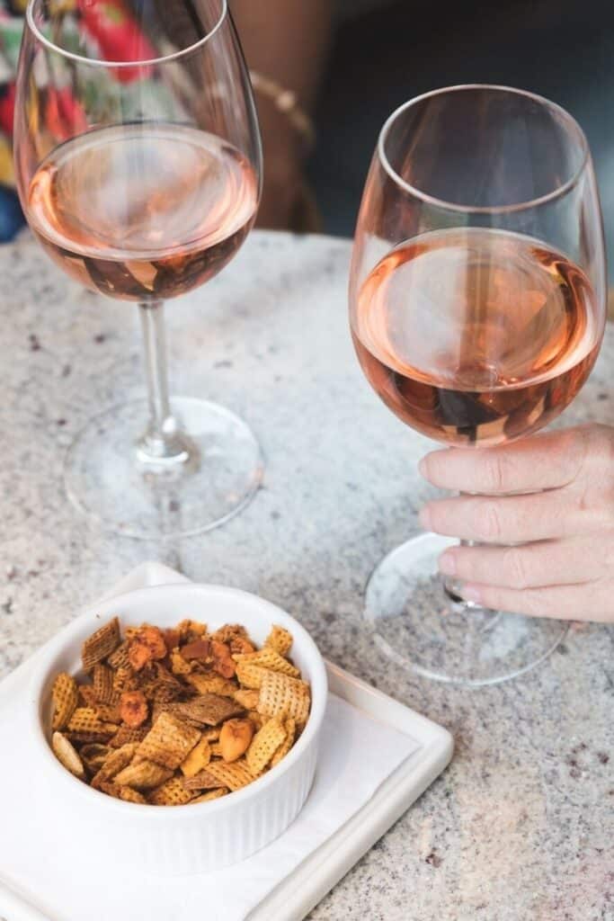 Two glasses of rosé wine on a stone tabletop, with a hand holding one glass. A white bowl of Chex Mix snacks sits nearby on a square white plate with a napkin.