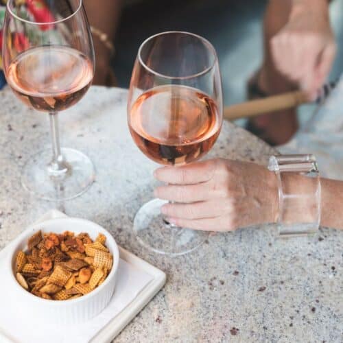 A close-up of two wine glasses filled with rosé on a speckled tabletop, accompanied by a white bowl of Chex Mix snacks. A hand with a clear bracelet is holding one wine glass.