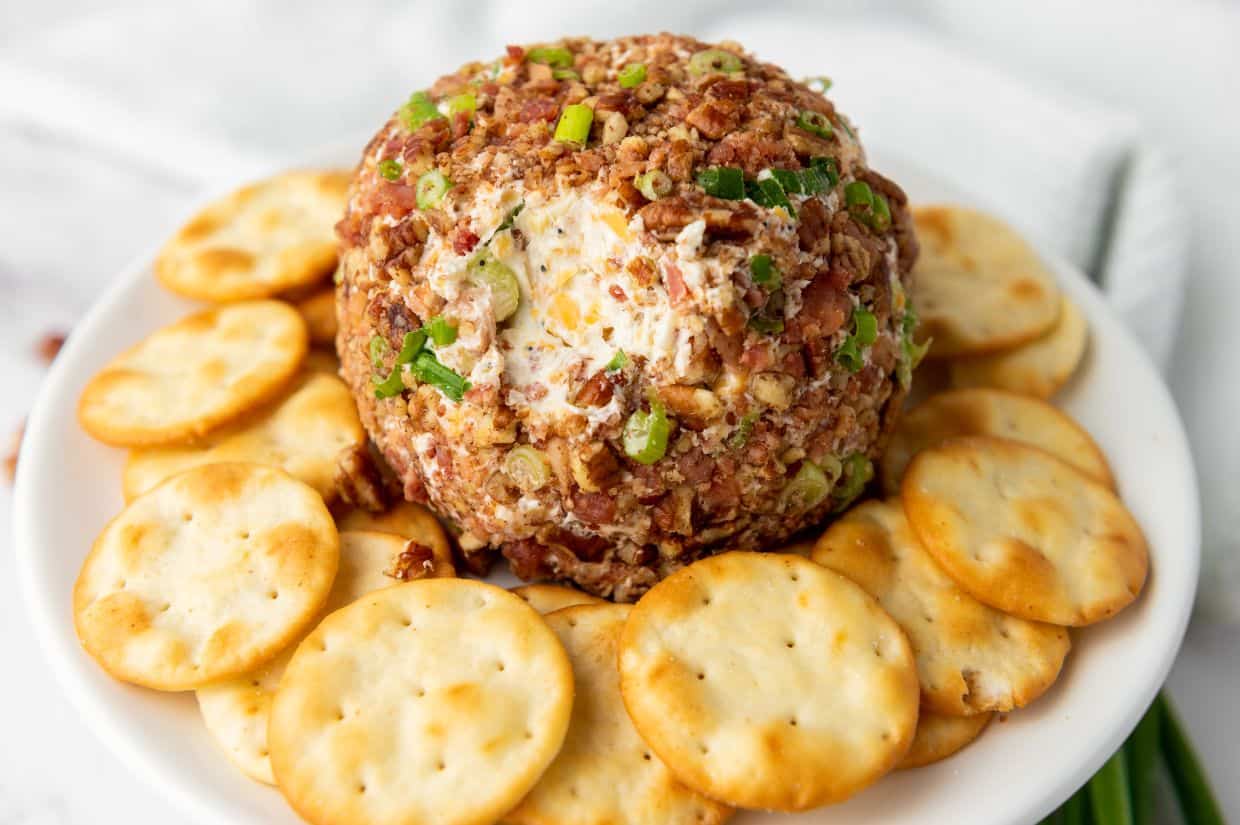 A hand holding a round cracker, reaching to dip into a pecan bacon cheeseball.