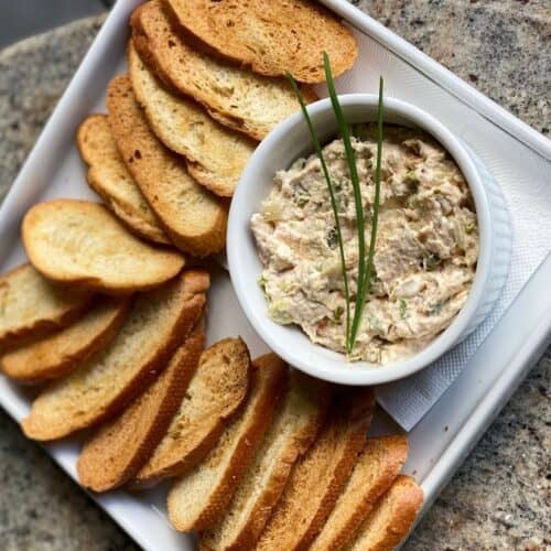 A bowl of creamy smoked trout dip garnished with fresh chives, served on a white tray alongside golden, crispy toasted bread slices.