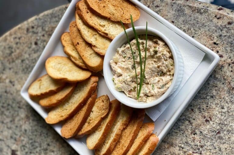 A bowl of creamy smoked trout dip garnished with fresh chives, served on a white tray alongside golden, crispy toasted bread slices.