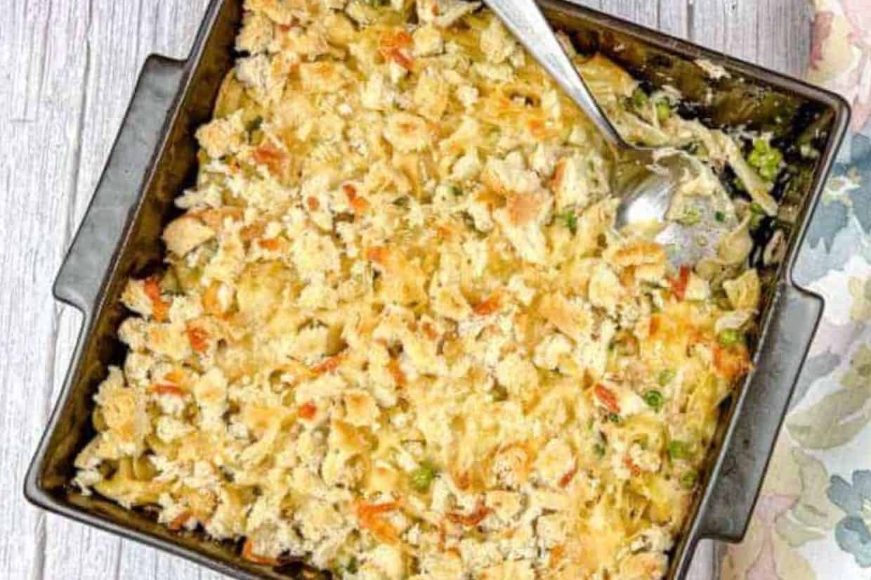 A baking dish of tuna noodle casserole topped with golden breadcrumbs, featuring creamy noodles, green peas, and a serving spoon on the side.