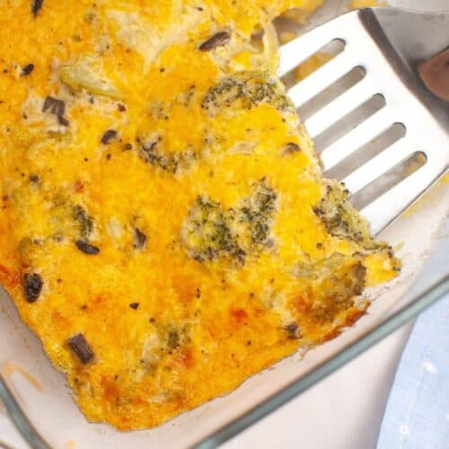 A close-up image of a golden-brown, baked broccoli cheese casserole in a glass dish. The dish features melted cheese with visible broccoli florets and small vegetable pieces, creating a textured surface.