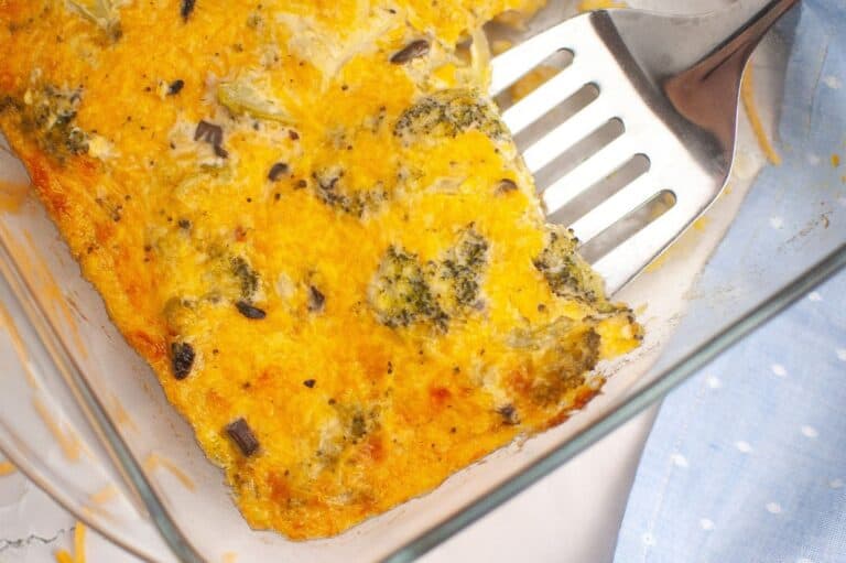 A close-up image of a golden-brown, baked broccoli cheese casserole in a glass dish. The dish features melted cheese with visible broccoli florets and small vegetable pieces, creating a textured surface.