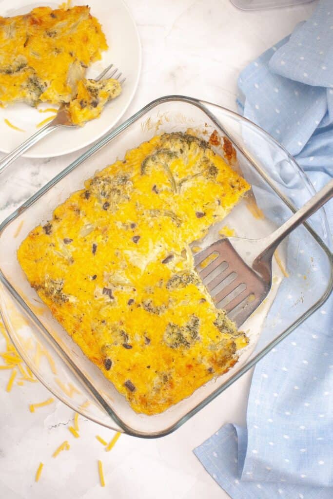 A glass baking dish with a golden-brown broccoli cheese casserole, partially served. A spatula rests inside the dish, and a serving is placed on a white plate in the background. 