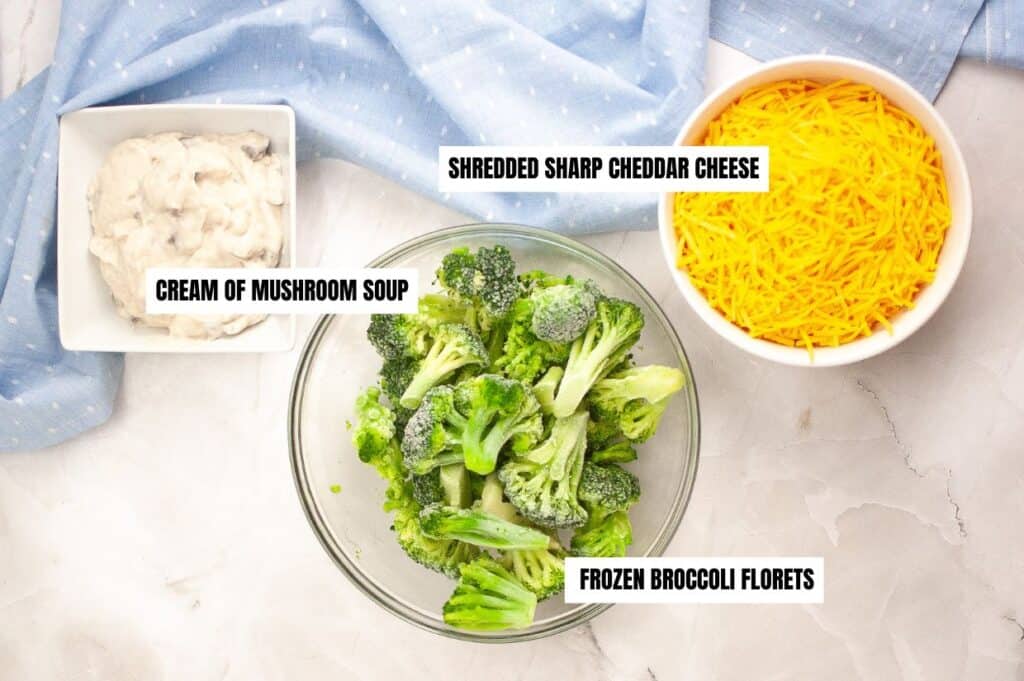 An image showing three ingredients for broccoli cheese casserole: cream of mushroom soup in a white bowl, frozen broccoli florets in a glass bowl, and shredded sharp cheddar cheese in a white bowl. 