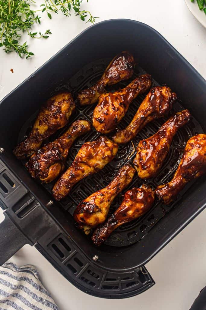 A batch of BBQ chicken drumsticks coated in a caramelized barbecue glaze sits in an air fryer basket. Fresh herbs and a striped kitchen towel are visible in the background, adding a touch of garnish and presentation.