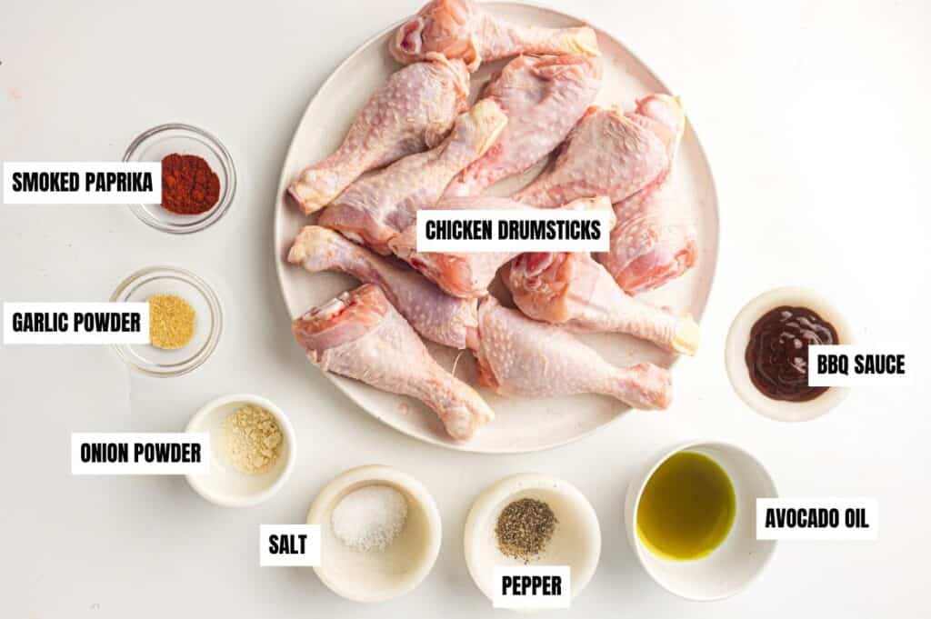 Ingredients for a BBQ chicken drumstick recipe are arranged on a white surface. The plate in the center contains raw chicken drumsticks. Surrounding it are small bowls labeled as smoked paprika, garlic powder, onion powder, salt, pepper, avocado oil, and BBQ sauce.