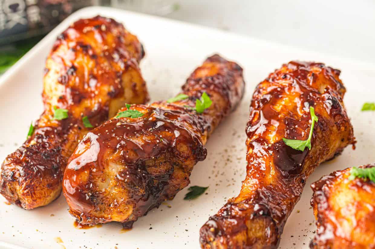 A close-up of crispy, air-fried BBQ chicken drumsticks glazed with a rich, glossy sauce and garnished with fresh parsley on a white plate.