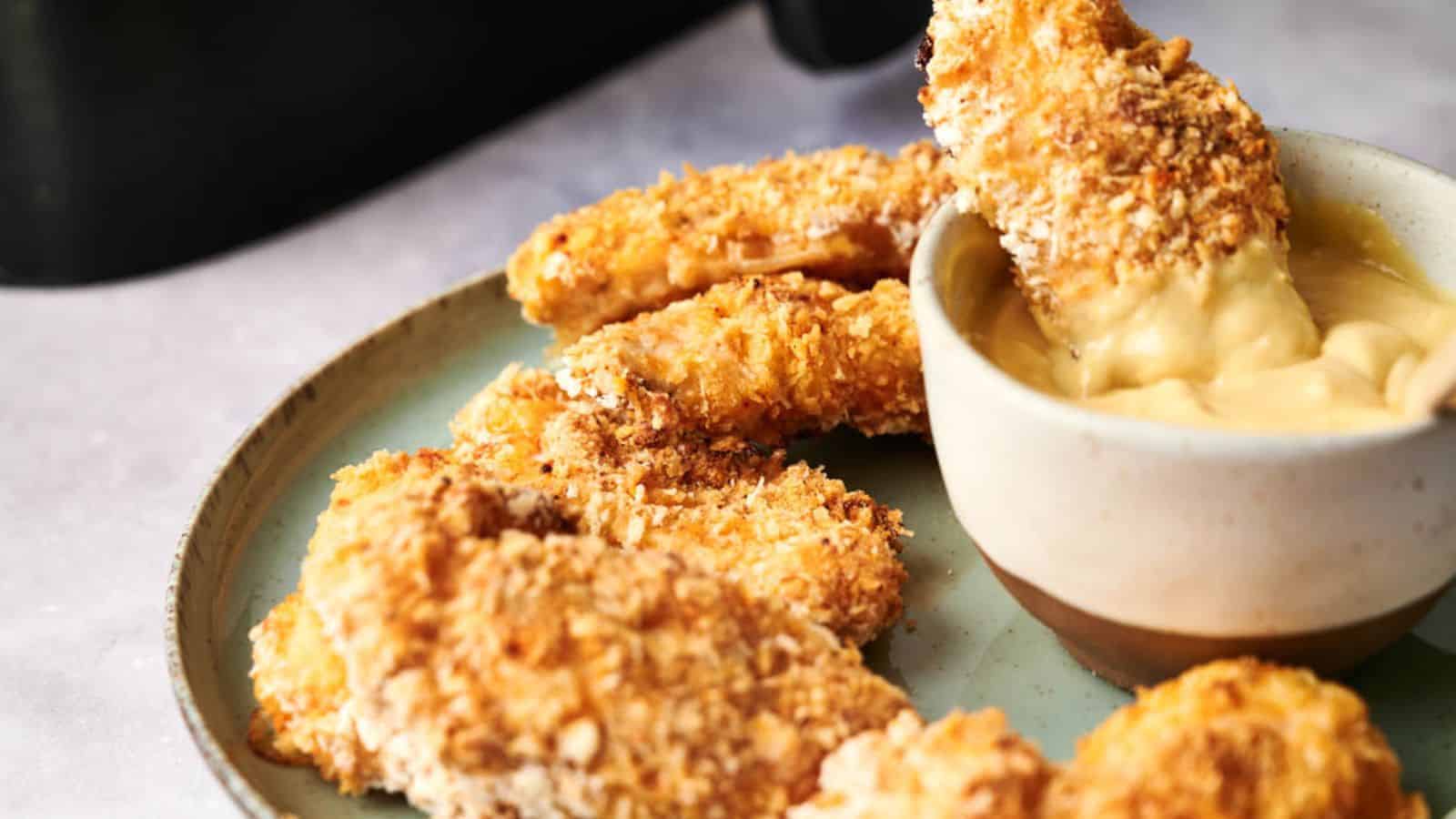 A plate of crispy Air Fryer Chicken Tenders next to a dipping bowl filled with creamy sauce.