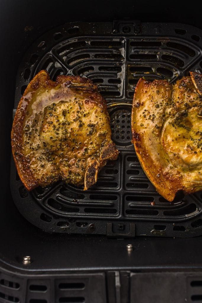 Two seasoned and cooked bone-in pork chops are displayed inside an air fryer basket. The chops are golden brown with visible pepper and herbs, glistening from their juices.