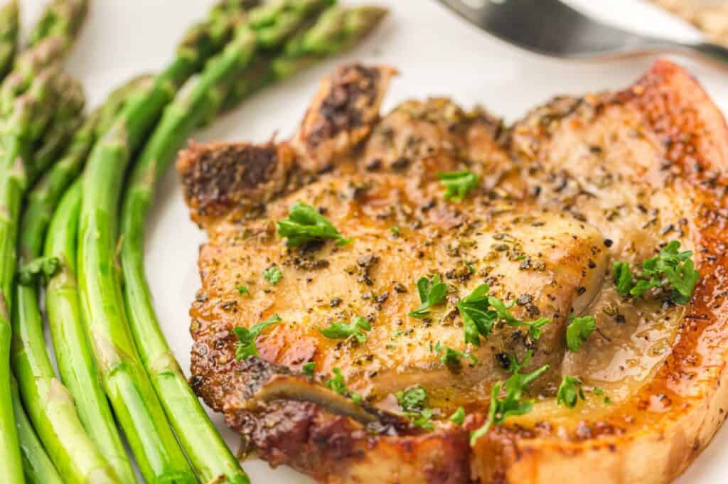 A close-up image of a golden-brown, seasoned pork chop garnished with fresh parsley on a white plate.