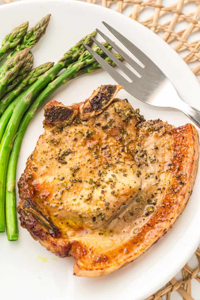 A golden brown, seasoned bone-in pork chop is served on a white plate alongside tender green asparagus spears.