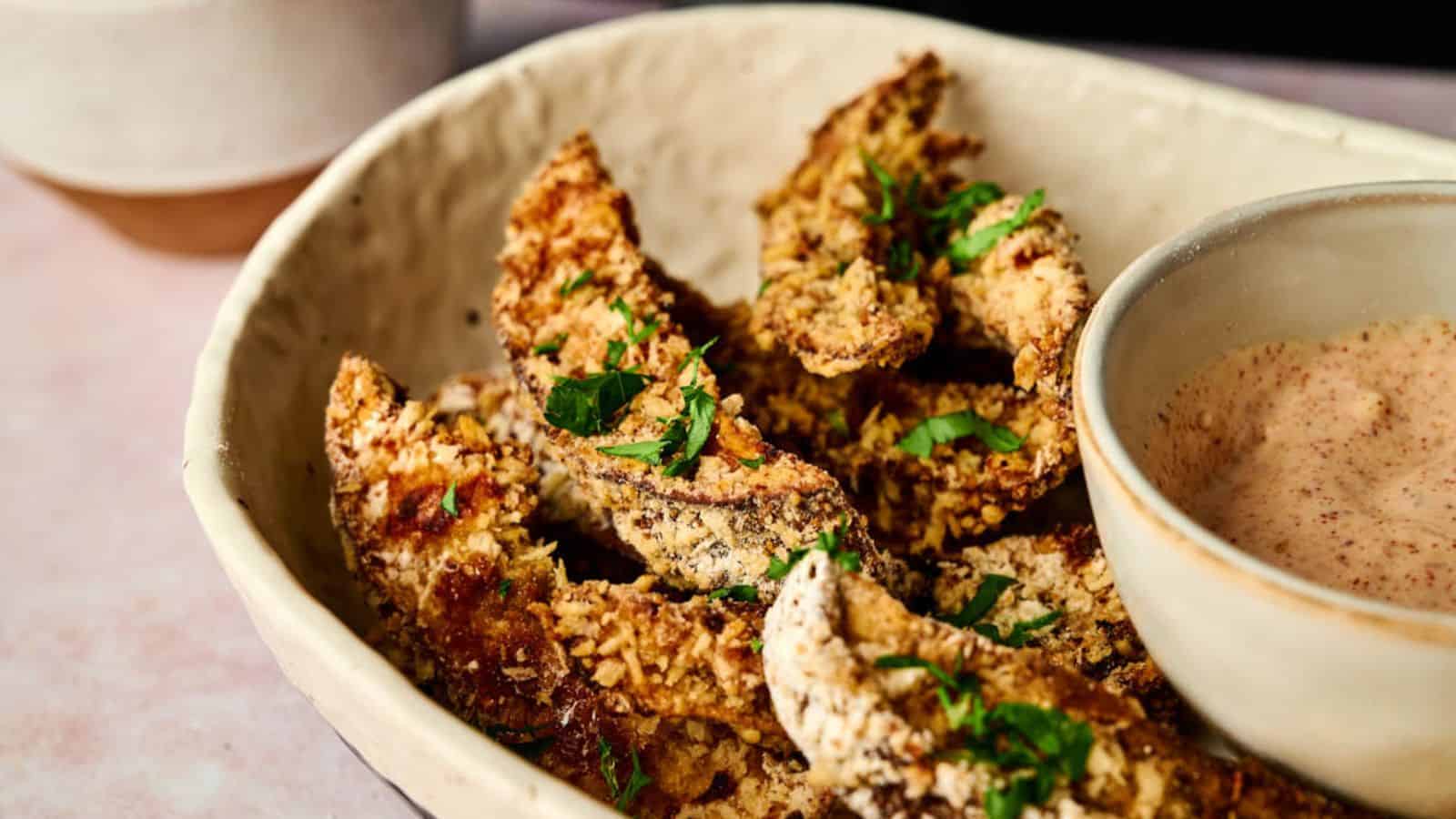 A bowl of crispy, breaded air fryer Portobello mushrooms garnished with fresh herbs, accompanied by a small bowl of dipping sauce.