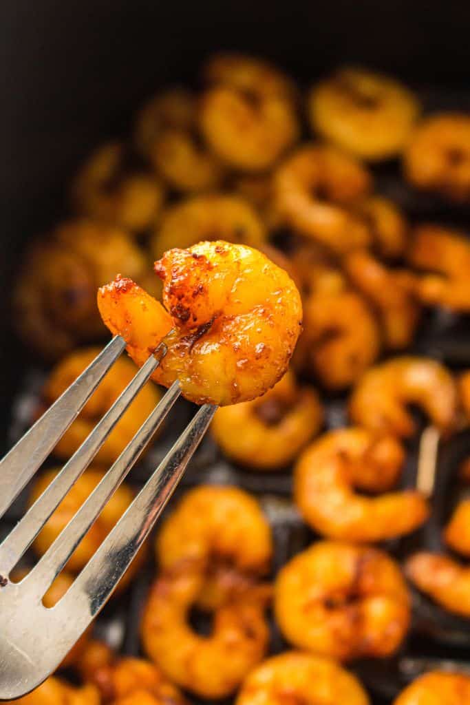 A close-up of a seasoned, golden-brown shrimp held on a fork, with more shrimp in the background cooked in an air fryer. The shrimp appear crispy and well-seasoned.