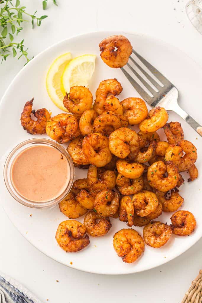 A white plate filled with crispy, seasoned air-fried shrimp, garnished with lemon slices and served with a creamy dipping sauce.