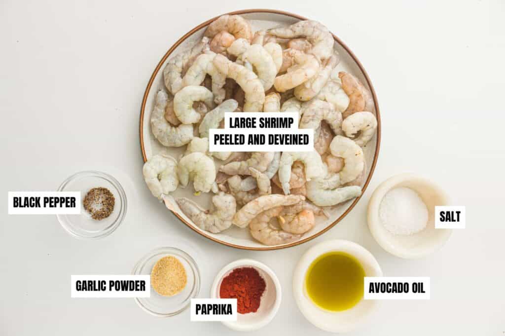 Ingredients for air fryer shrimp displayed on a surface: a plate of large peeled and deveined shrimp, bowls of black pepper, garlic powder, paprika, salt, and avocado oil.