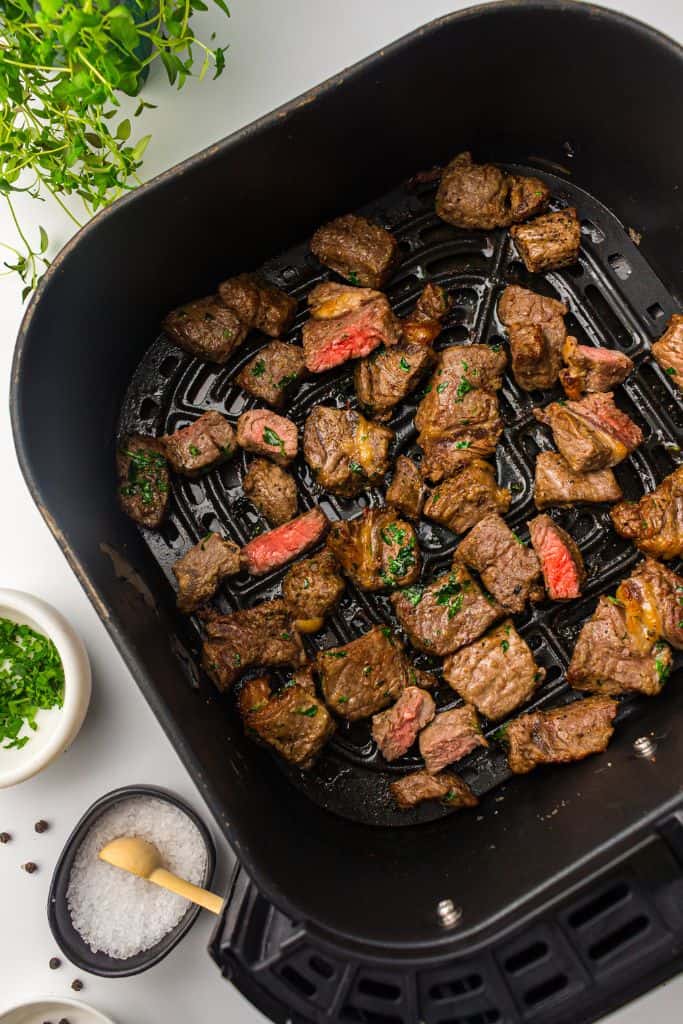 Cooked steak bites inside an air fryer basket, beautifully browned and garnished with chopped herbs.