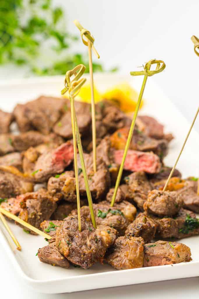 A plate of juicy, bite-sized steak pieces seasoned with herbs and spices, served with skewers for easy picking.