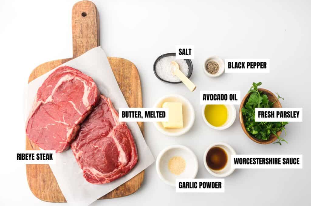 Ingredients for Air Fryer Steak Bites neatly arranged on a white surface. Two raw ribeye steaks are placed on a wooden cutting board. Surrounding the board are small bowls containing salt, black pepper, melted butter, avocado oil, Worcestershire sauce, garlic powder, and fresh parsley.