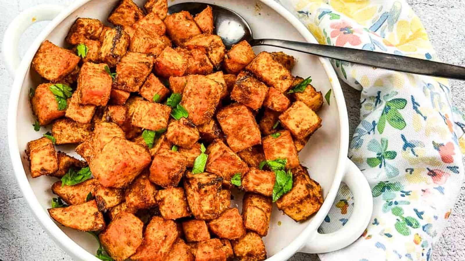 A white bowl filled with seasoned roasted sweet potato cubes, garnished with chopped herbs, with a metal serving spoon on the side and a printed napkin partially visible.