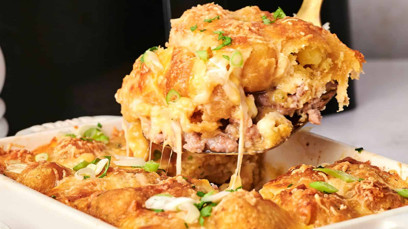 A slice of cheesy casserole with ground meat is being lifted from a baking dish, topped with chopped green onions.