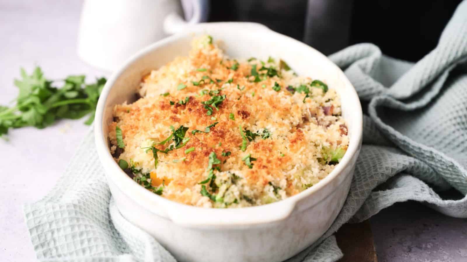 A white ceramic dish containing baked vegetable gratin topped with browned breadcrumbs and parsley, placed on a light green textured cloth.